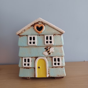 May include: A light blue ceramic house with a yellow door, white windows, and a bee design. The house has a heart-shaped window on the top.