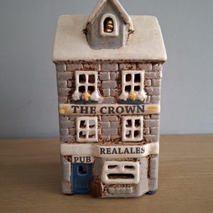 以下が含まれることがあります： 「THE CROWN」と「REAL ALES」の文字が入った、パブの形をした陶器製の置物。灰色のレンガ造りの外観、白いトリム、青いドアが特徴です。屋根は白く、小さなドーマーウィンドウが付いています。