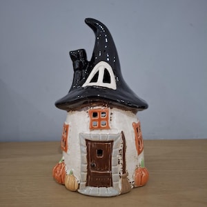 May include: A ceramic house-shaped figurine featuring a tall, black witch hat roof with a small window. The off-white structure has a brown door, orange-trimmed windows, and orange pumpkins at the base. A broom is sculpted next to the front door.