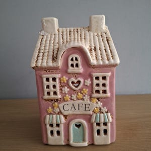 May include: A pink ceramic cafe ornament with a white roof and chimney. The word "CAFE" is displayed above the door. The facade is decorated with windows, flowers, and a heart-shaped cutout.