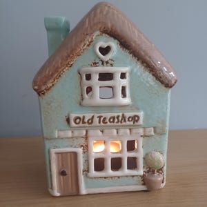 May include: A ceramic teal-colored house-shaped candle holder with a brown thatched roof. The words "Old Teashop" are above the window. A small heart-shaped cutout is on the roof. A small potted plant is on the side.