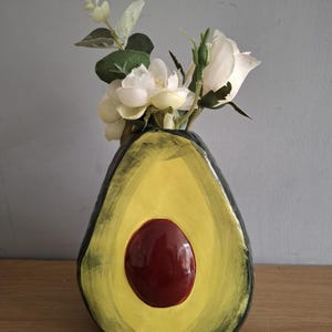 Op de afbeelding: Een keramische vaas in de vorm van een avocado, met een gele binnenkant en een donkerrode pit. De buitenkant is groen en zwart geschilderd. De vaas bevat witte bloemen en groene bladeren, wat een uniek decoratief element creëert.