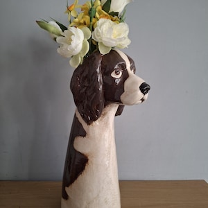 Könnte beinhalten: Keramikvase in Form eines Hundekopfes mit braunen und weißen Markierungen. Die Vase ist mit künstlichen weißen und gelben Blumen gefüllt. Der Hundekopf ist detailliert mit braunen Ohren, einer weißen Schnauze und braunen Augen.