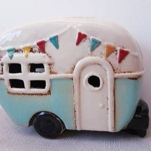 Può includere: Figurina a forma di camper in ceramica bianca, azzurra e marrone, con una ruota nera. Il camper ha una porta, una finestra e una ghirlanda di bandierine colorate. Un oggetto decorativo.
