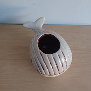 May include: A ceramic whale-shaped planter in a light gray color with brown accents. The whale has a textured, ribbed design and a dark interior. The tail fin is upright.