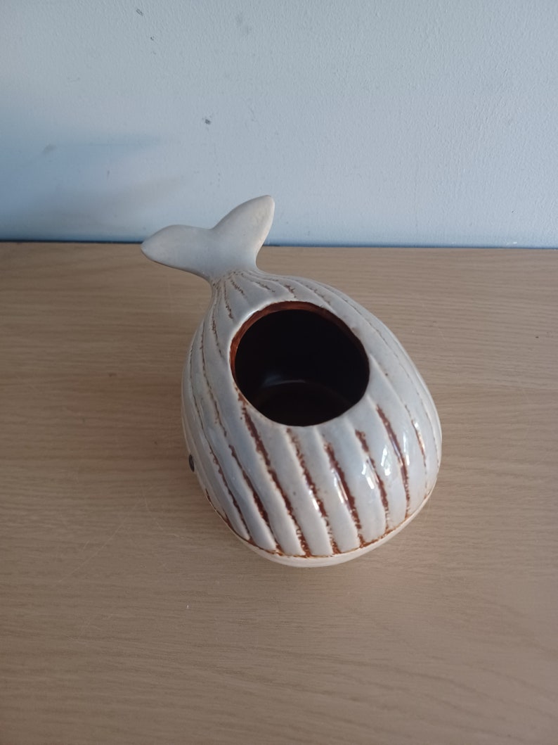 May include: A ceramic whale-shaped planter in a light gray color with brown accents. The whale has a textured, ribbed design and a dark interior. The tail fin is upright.
