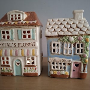 May include: Two ceramic house-shaped candle holders. One is white with the text "Petal's Florist" and a brown roof. The other is blue with the text "Village Store" and a white roof. Both have windows and doors.