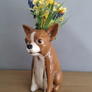 May include: A ceramic dog vase in shades of brown and white, filled with yellow and blue flowers. The vase is on a wooden surface. The dog-shaped vase has a glossy finish and a white stripe on its head.