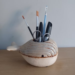 May include: A ceramic whale-shaped desk organizer in shades of cream and gray, holding various art supplies. The organizer features a brown rim and a small, dark eye. Items inside include paintbrushes, pens, and scissors.