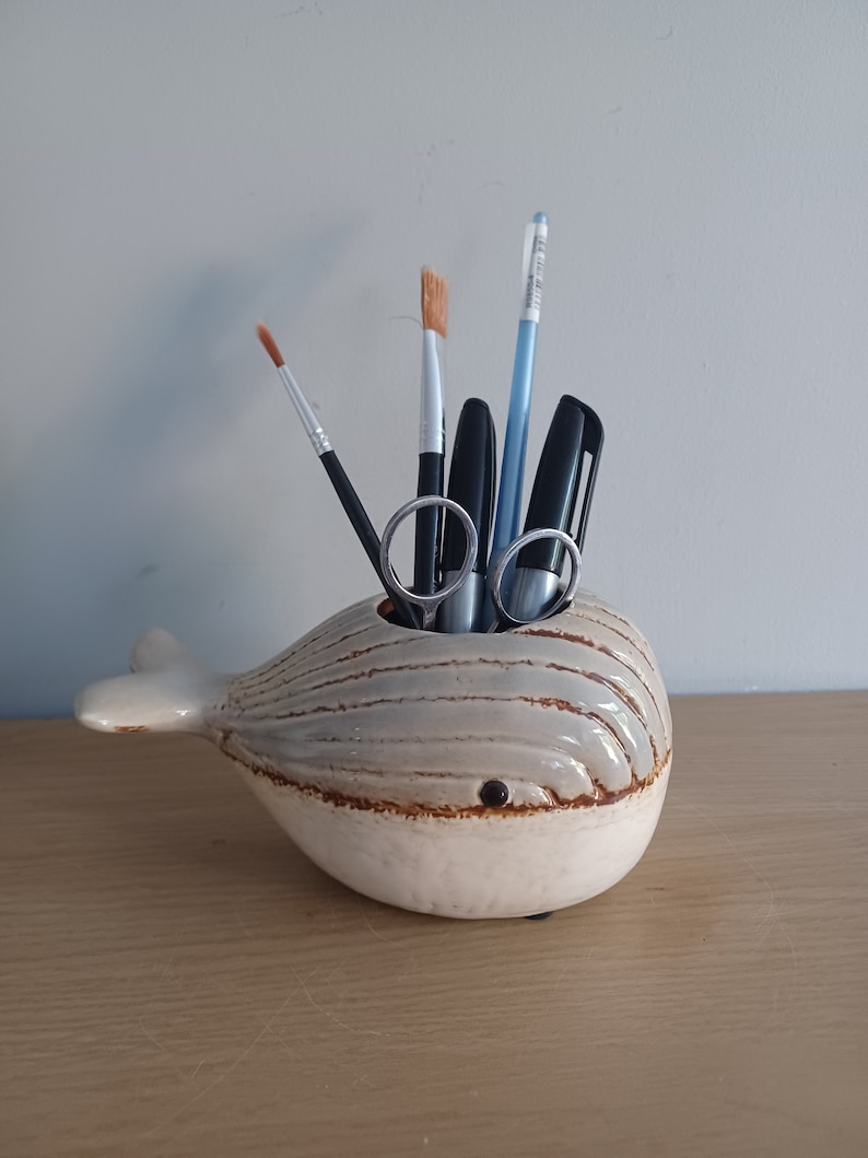 May include: A ceramic whale-shaped desk organizer in shades of cream and gray, holding various art supplies. The organizer features a brown rim and a small, dark eye. Items inside include paintbrushes, pens, and scissors.