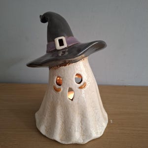 May include: A ceramic ghost figurine wearing a dark grey witch hat with a purple band and a white buckle. The ghost has a cream-coloured, textured body with cutouts for eyes and a mouth, illuminated from within by a warm, glowing light source.