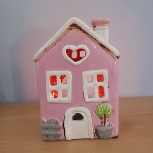 May include: A pink ceramic house with a white chimney, a heart-shaped window, and two windows with white frames. The house has a white door with a black knob and a small green bush in a grey pot.