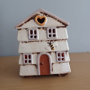 May include: A white ceramic house with a brown roof and a bee design. The house has a heart-shaped window and a brown door.