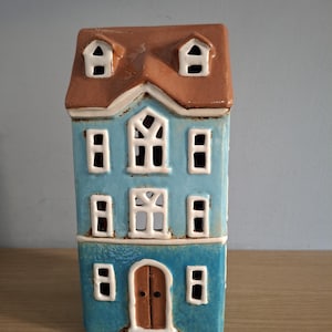 May include: A blue ceramic house-shaped candle holder with a brown roof and white window frames. The house has a brown front door and multiple windows. The candle holder is designed to hold a tea light. The measurements are in centimetres.