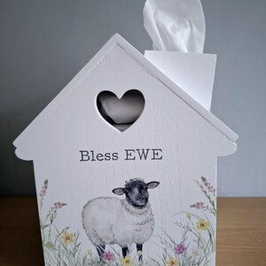 Op de afbeelding: Witte houten tissueboxhoes in de vorm van een huis, met een hartvormige uitsparing en de tekst "Bless EWE". Een aquarel illustratie van een schaap staat op de voorkant, omringd door wilde bloemen. Witte tissues zijn zichtbaar.