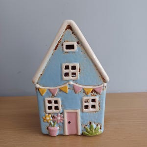 May include: A ceramic house ornament with a light blue exterior, white roof, and pink door. The house features small square windows, a string of colourful pennant flags, and decorative flowers in a pot. The house is approximately 15 cm tall.