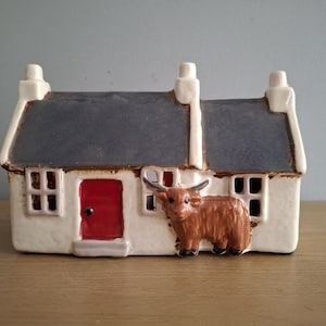 Op de afbeelding: Een keramieken huisje met een donkergrijs dak, witte muren en een rode deur. Een bruin Highland-koeienbeeldje is aan de voorkant bevestigd. Het huisje heeft twee schoorstenen en twee ramen.