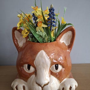 May include: A ceramic cat-shaped planter in shades of brown and white, filled with yellow and blue artificial flowers and greenery. The planter has a detailed face with large eyes and paws. A decorative item for home decor.