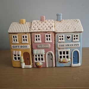 May include: A ceramic decorative piece depicting three colorful buildings. The buildings are yellow, pink, and blue, with white roofs and windows. The yellow building says "GIFT BOX", the pink building says "Tea Rooms", and the blue building says "THE SWAN INN".