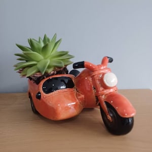 May include: A ceramic planter shaped like a vintage motorcycle with a sidecar, painted in a glossy orange. The sidecar holds a green succulent plant. The motorcycle has black tires and handlebars, and a white headlight.