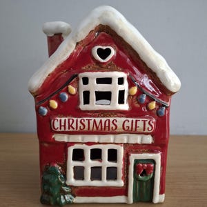 Può includere: Una statuetta a forma di casa in ceramica rossa con tetto e camino coperti di neve bianca. La parte anteriore presenta un'apertura a forma di cuore, finestre, una porta verde con una ghirlanda, un piccolo albero e la scritta CHRISTMAS GIFTS.