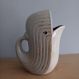 Può includere: Un'anfora in ceramica bianca a forma di balena con una superficie testurizzata e un occhio marrone.