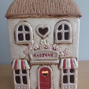 May include: A ceramic house-shaped candle holder with a thatched roof. The off-white building has arched windows, a heart-shaped opening, and a red door. The word "ANTIQUE" is displayed above the door.
