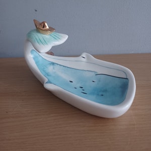 May include: A ceramic whale-shaped dish with a blue watercolour design and a gold-coloured hat. The dish is white with a curved shape, ideal for holding small items. The whale has a light blue and green accent on its head.