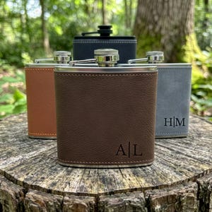 May include: Four metal flasks wrapped in leather-like material in brown, tan, grey, and black. The front-most brown flask features the engraved initials A|L, while the grey flask displays H|M. Each flask has a silver-toned screw-top cap and metal rim.