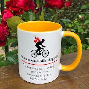May include: A white ceramic mug with a yellow rim and handle. The mug features a black silhouette of a person riding a bicycle with flames coming from the back wheel. The text on the mug reads "Being an engineer is like riding a bike *Except the bike is on fire You're on fire Everything is on fire And Your In Hell"