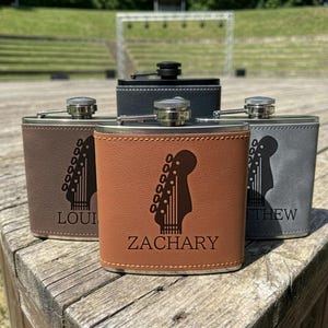 May include: Four metal flasks wrapped in faux leather in shades of brown, tan, and grey. Each flask features a laser-engraved graphic of a guitar headstock and a name, including Zachary, Louis, and Matthew, with a silver screw-top cap.