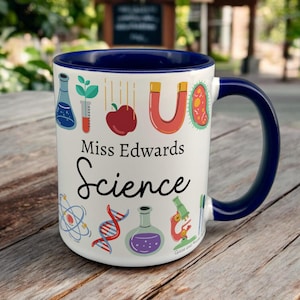 May include: White ceramic coffee mug with a blue rim and handle. The mug has a science theme with illustrations of beakers, test tubes, a microscope, a DNA strand, and an apple. The text "Miss Edwards Science" is printed on the mug.