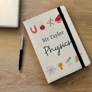 May include: A beige canvas notebook with a black elastic closure. The notebook has a personalised inscription that reads "Mr Taylor Physics" and is decorated with colourful science-themed illustrations, including an atom, a light bulb, a telescope, a planet, a magnet, an apple, and a molecule.
