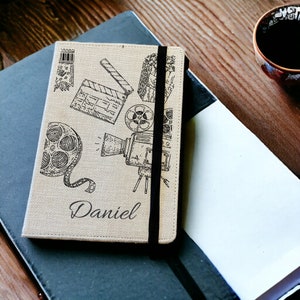 May include: A beige notebook featuring film-related illustrations such as a clapperboard and film reel, with the name "Daniel" printed on the cover. It has a black elastic band. The notebook rests on a dark grey notebook and a white notepad.