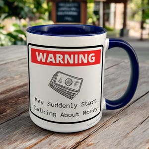 May include: White ceramic mug with a navy blue interior and handle. The mug features a red and white warning label with the text "WARNING" and a graphic of money. Below the graphic, the text reads "May Suddenly Start Talking About Money."