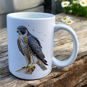 White Ceramic Mug with Falcon Design sat on a table. The Cup has a standard handle. Customisable text is printed below the Falcon design in a black text.