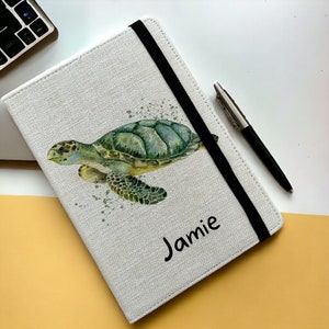 May include: A white canvas notebook with a black elastic closure. The cover features a watercolour illustration of a sea turtle with green and blue shells. The name "Jamie" is printed in black below the turtle.