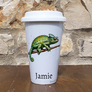 May include: White ceramic travel mug featuring a green chameleon illustration and the name "Jamie" printed on the bottom. The mug has a lid.