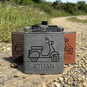 May include: Three rectangular metal flasks wrapped in faux leather in grey, brown, and tan. Each flask features a laser-engraved scooter graphic and a personalised name. The front grey flask is clearly engraved with the name ETHAN below the scooter.