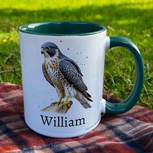 Green & White Ceramic Mug with Falcon Design sat on a table. The Cup has a standard handle. Customisable text is printed below the Falcon design in a black text.
