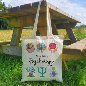 Puede incluir: Una bolsa de lona blanca con un tema de psicolog&iacute;a. La bolsa tiene ilustraciones coloridas de cerebros, una pieza de rompecabezas, un &aacute;rbol y un reloj. El texto "Mrs May Psychology" y una letra griega Psi verde est&aacute;n impresos en la bolsa.