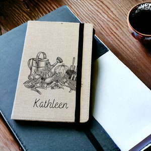 May include: A beige linen journal with a black elastic closure. The cover features a black and white illustration of gardening tools and produce, including a watering can, a pumpkin, and a trowel. The name "Kathleen" is printed in black below the illustration.