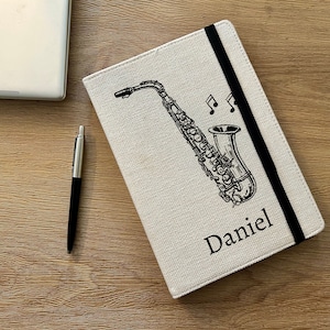 May include: A beige linen notebook with a black elastic closure and a black and white illustration of a saxophone with musical notes. The name "Daniel" is printed on the front of the notebook.