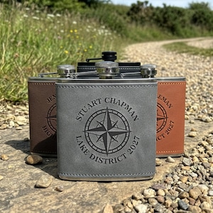 May include: Three rectangular metal flasks wrapped in leather-like material in grey, dark brown, and tan. Each flask features a screw-top lid and a laser-engraved compass design with the text STUART CHAPMAN LAKE DISTRICT 2027 centred on the front.