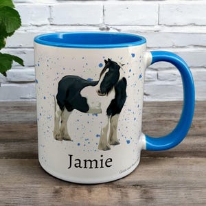 Blue & White Ceramic Mug with Horse Design sat on a table. The Cup has a standard handle. Customisable text is printed below the Horse design in a black text.