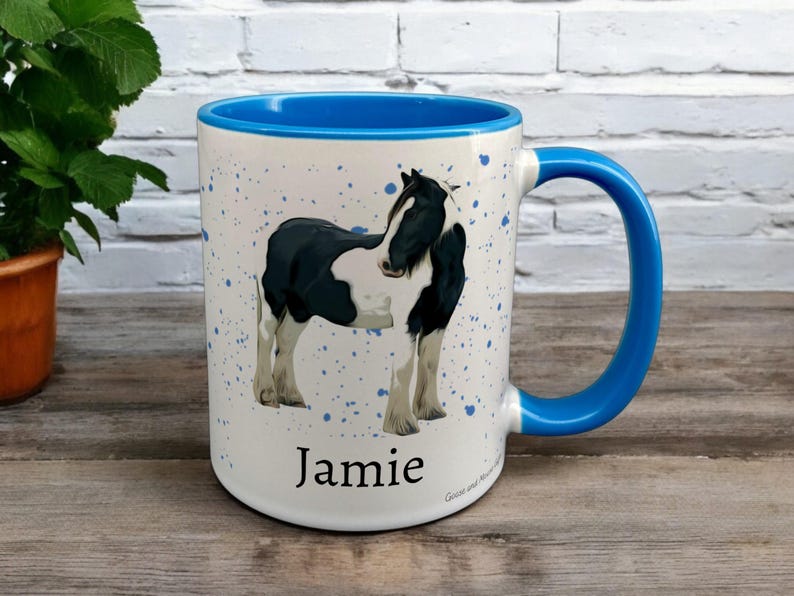 Blue & White Ceramic Mug with Horse Design sat on a table. The Cup has a standard handle. Customisable text is printed below the Horse design in a black text.