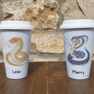 May include: Two white travel mugs with lids. The mug on the left has a yellow snake design and the name "Leila" printed on it. The mug on the right has a grey snake design and the name "Harry" printed on it.