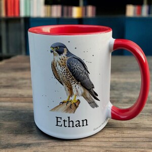 Red & White Ceramic Mug with Falcon Design sat on a table. The Cup has a standard handle. Customisable text is printed below the Falcon design in a black text.