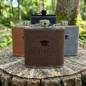 May include: A collection of four rectangular metal flasks wrapped in faux leather in brown, tan, and grey colours. Each flask features an engraved graduation cap icon, the name Bethany, and the text Graduation Class of 2027 on the front surface.