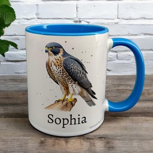 Blue & White Ceramic Mug with Falcon Design sat on a table. The Cup has a standard handle. Customisable text is printed below the Falcon design in a black text.